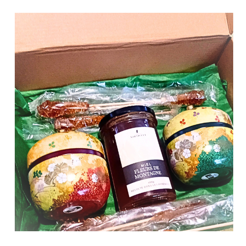 Coffret ouvert contenant deux boîtes à thé décoratives, un pot de miel de fleurs de montagne, et plusieurs bâtonnets de sucre candi, sur fond de papier vert