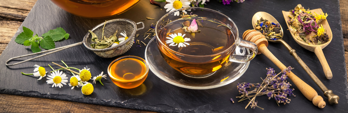 Infusion à base de plantes et fleurs servie dans une tasse en verre, accompagnée d’une théière transparente, d’un filet de miel, de fleurs fraîches et de cuillères remplies de tisanes séchées, le tout posé sur une ardoise noire dans une atmosphère chaleureuse et naturelle.